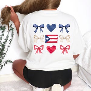 May include: White t-shirt featuring a design of blue bows, a blue heart, tan bows, the Puerto Rican flag, red bows, and a red heart. The design is centered on the back of the shirt. The shirt is a casual, comfortable style.