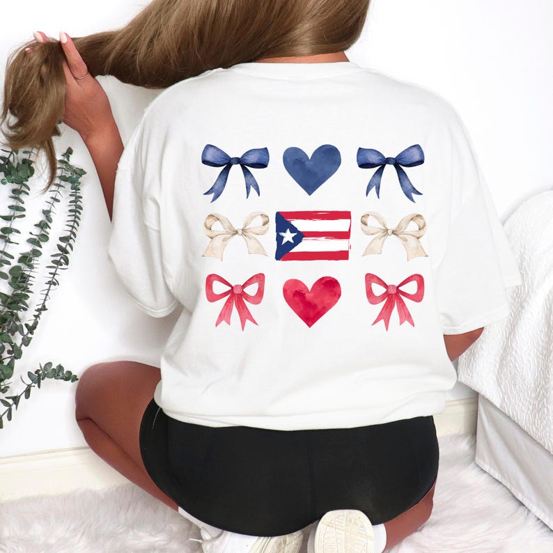May include: White t-shirt featuring a design of blue bows, a blue heart, tan bows, the Puerto Rican flag, red bows, and a red heart. The design is centered on the back of the shirt. The shirt is a casual, comfortable style.