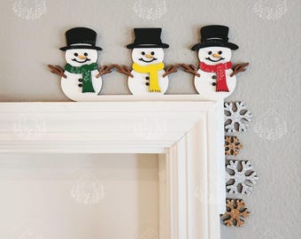 May include: Three snowman decorations with black top hats, brown stick arms, and colorful scarves in green, yellow, and red. The snowmen are placed above a white door frame, with snowflake decorations on the wall.