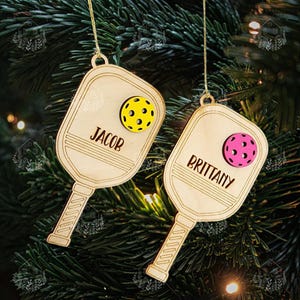 May include: Two wooden pickleball paddle ornaments with the names "Jacob" and "Brittany" on them. Each ornament has a small yellow or pink pickleball attached to it.