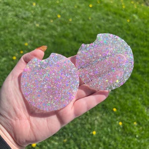 May include: Two round car coasters with a glittery pink and iridescent design. Each coaster has a small notch for easy removal from a car cup holder. The coasters are held in a hand against a background of green grass.