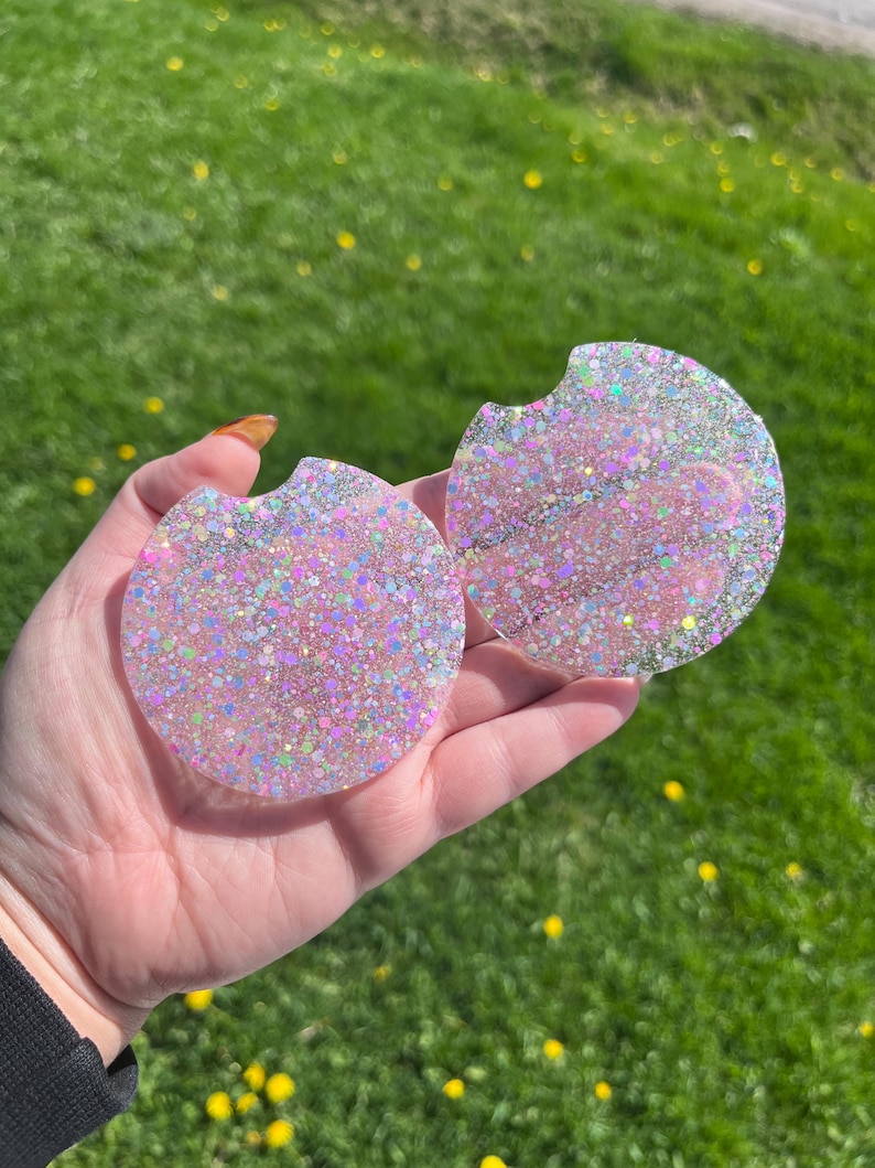 Op de afbeelding: Twee ronde auto-onderzetters met een glitterroze en iriserend ontwerp. Elke onderzetter heeft een kleine inkeping voor gemakkelijk verwijderen uit een bekerhouder. De onderzetters worden in een hand gehouden tegen een achtergrond van groen gras.