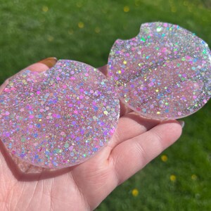 Op de afbeelding: Twee ronde auto-onderzetters met een glitterende, iriserende afwerking. De onderzetters zijn roze met een mix van kleurrijke glitter, wat een sprankelend effect cre&euml;ert. E&eacute;n onderzetter heeft een kleine inkeping.