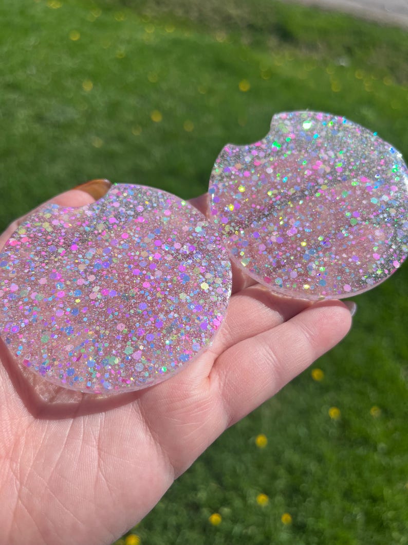 Op de afbeelding: Twee ronde auto-onderzetters met een glitterende, iriserende afwerking. De onderzetters zijn roze met een mix van kleurrijke glitter, wat een sprankelend effect cre&euml;ert. E&eacute;n onderzetter heeft een kleine inkeping.