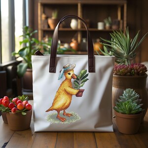 Pode incluir: Sacola de lona branca com alças de couro marrom. A sacola apresenta uma imagem bordada colorida de um pato amarelo usando um chapéu azul e branco segurando uma planta em vaso.
