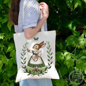Pode incluir: Uma bolsa tote de lona branca com um coelho verde e marrom vestindo um vestido verde e segurando um pincel. O coelho está rodeado de folhas verdes e uma borboleta. O texto "Kvitko Mania Stitch" é visível no canto inferior direito da imagem.