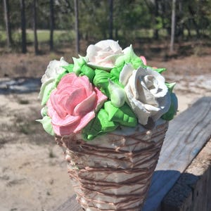May include: A decorative basket filled with colorful, handcrafted flowers. The arrangement features pink, white, and cream-colored roses with green leaves. The basket has a woven design with brown accents, set against a natural outdoor backdrop.