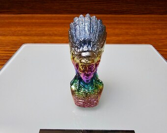 May include: A small, rainbow-colored metal sculpture of a person with a feathered headdress. The sculpture is on a white digital scale that reads 77 grams.