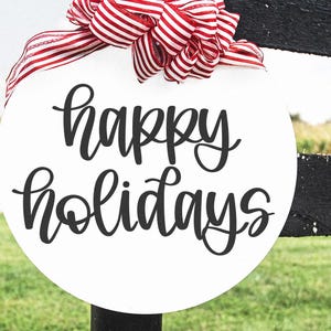 K&ouml;nnte beinhalten: Ein wei&szlig;es, rundes Schild mit den Worten "happy holidays" in schwarzer Schrift. Oben ist eine rot-wei&szlig; gestreifte Schleife befestigt. Das Schild h&auml;ngt an einem schwarzen Zaunpfosten mit gr&uuml;nem Gras im Hintergrund.