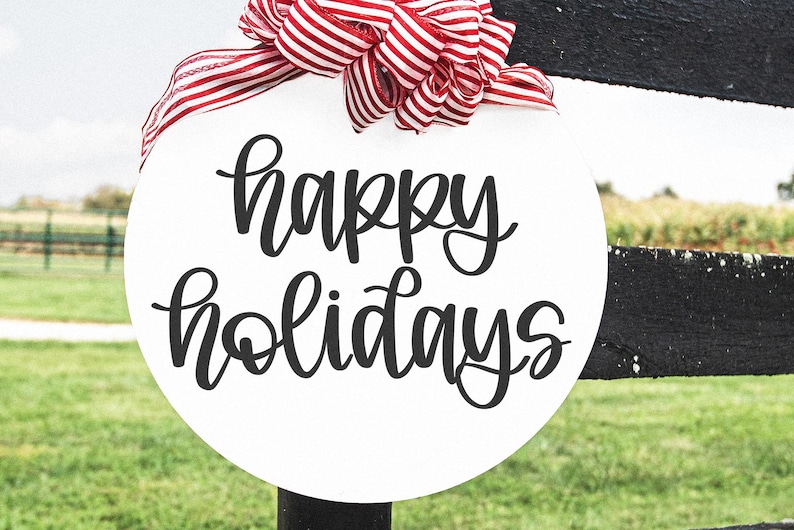 K&ouml;nnte beinhalten: Ein wei&szlig;es, rundes Schild mit den Worten "happy holidays" in schwarzer Schrift. Oben ist eine rot-wei&szlig; gestreifte Schleife befestigt. Das Schild h&auml;ngt an einem schwarzen Zaunpfosten mit gr&uuml;nem Gras im Hintergrund.