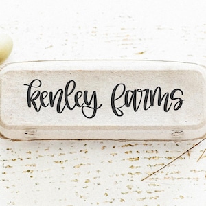 K&ouml;nnte beinhalten: Eine rustikale Eierschachtel mit dem Schriftzug "Kenley Farms" in eleganter Schrift. Zwei Eier, eines hellgr&uuml;n und eines hellbraun, liegen neben der Schachtel. Getrocknete Weizenhalme verleihen der Komposition eine nat&uuml;rliche Note, alles auf einer wei&szlig;en Holzoberfl&auml;che.