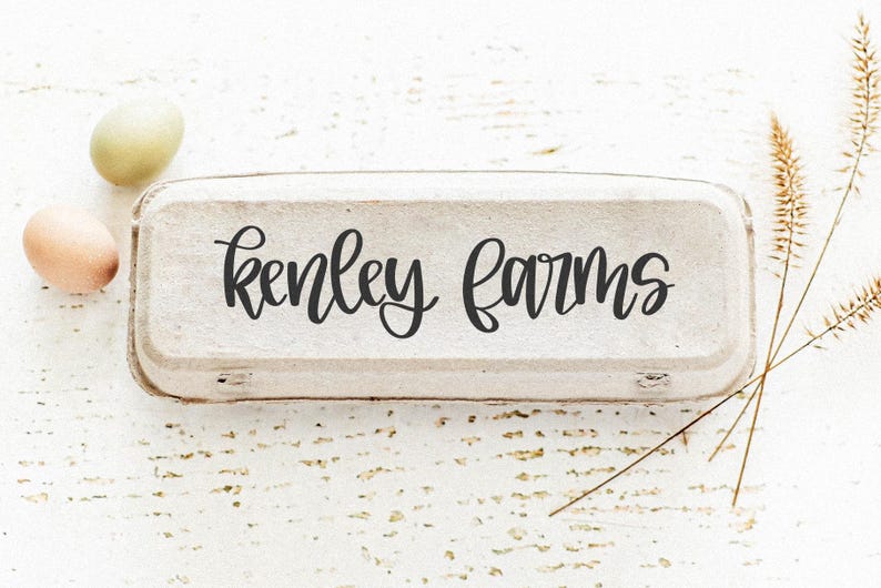 K&ouml;nnte beinhalten: Eine rustikale Eierschachtel mit dem Schriftzug "Kenley Farms" in eleganter Schrift. Zwei Eier, eines hellgr&uuml;n und eines hellbraun, liegen neben der Schachtel. Getrocknete Weizenhalme verleihen der Komposition eine nat&uuml;rliche Note, alles auf einer wei&szlig;en Holzoberfl&auml;che.