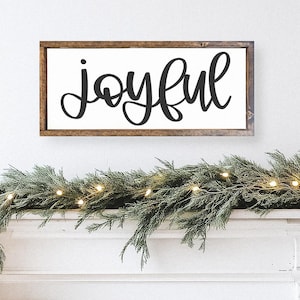 K&ouml;nnte beinhalten: Rechteckiges Holzschild mit dem Wort "joyful" in schwarzer Schreibschrift. Das Schild ist an einer wei&szlig;en Wand &uuml;ber einer Girlande aus Gr&uuml;n und Lichtern angebracht und schafft ein festliches Urlaubs-Thema.