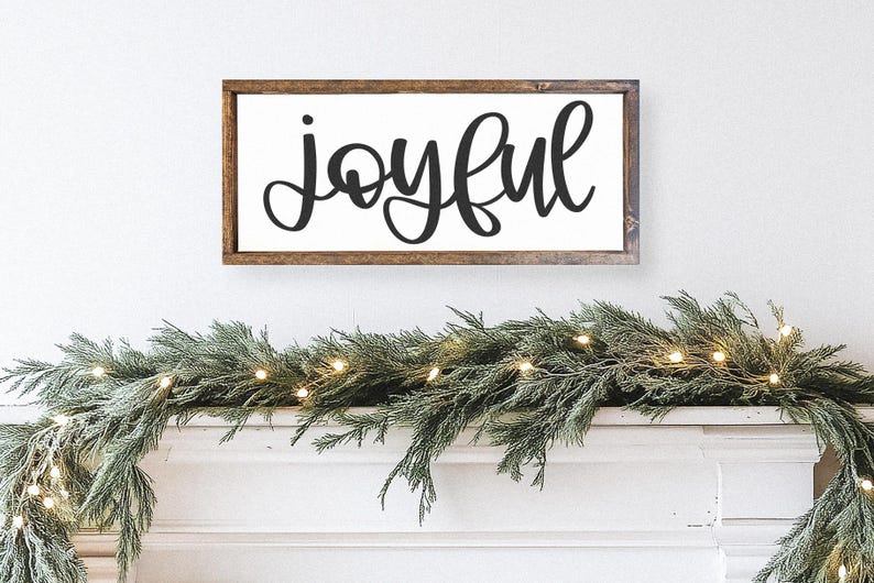 K&ouml;nnte beinhalten: Rechteckiges Holzschild mit dem Wort "joyful" in schwarzer Schreibschrift. Das Schild ist an einer wei&szlig;en Wand &uuml;ber einer Girlande aus Gr&uuml;n und Lichtern angebracht und schafft ein festliches Urlaubs-Thema.