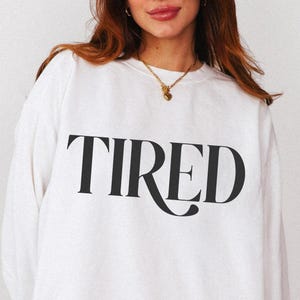 May include: White sweatshirt with the word "TIRED" printed in large, black, stylized letters across the front. The sweatshirt has long sleeves and a crew neck. The overall style is casual and comfortable.