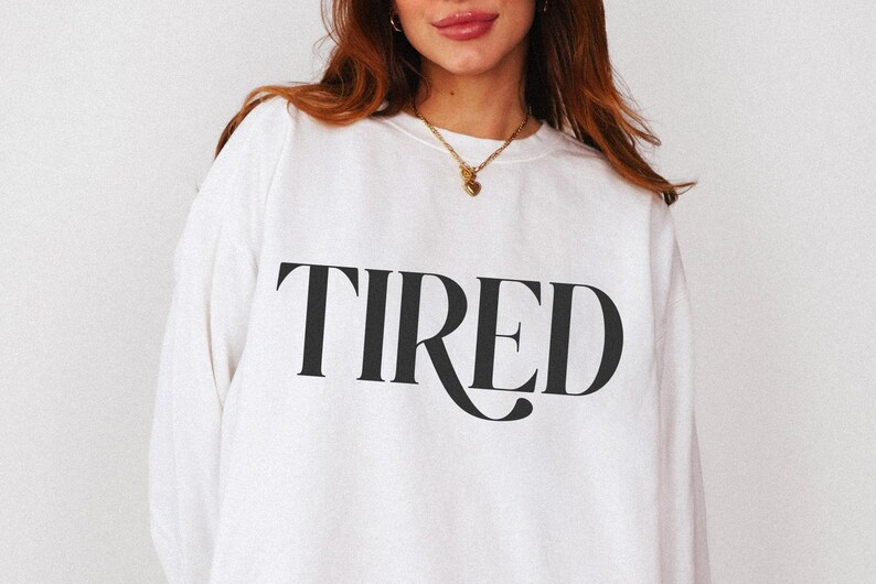 May include: White sweatshirt with the word "TIRED" printed in large, black, stylized letters across the front. The sweatshirt has long sleeves and a crew neck. The overall style is casual and comfortable.