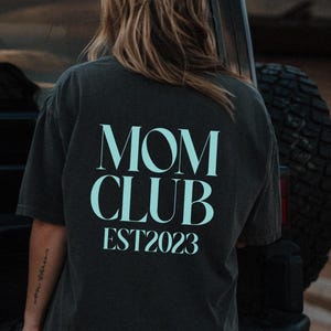 May include: Dark gray t-shirt with the words "MOM CLUB EST2023" printed in a light blue color. The shirt is worn by a person standing near a black vehicle.