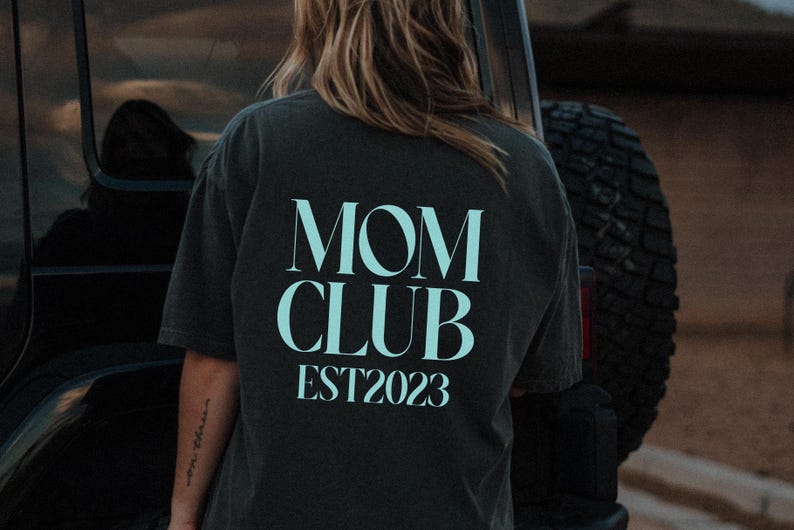 May include: Dark gray t-shirt with the words "MOM CLUB EST2023" printed in a light blue color. The shirt is worn by a person standing near a black vehicle.
