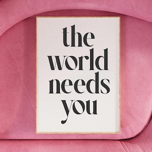 May include: A framed print with the words "the world needs you" in black, elegant typography. The print is set against a white background and framed in a light wood. The background is a pink, textured surface.