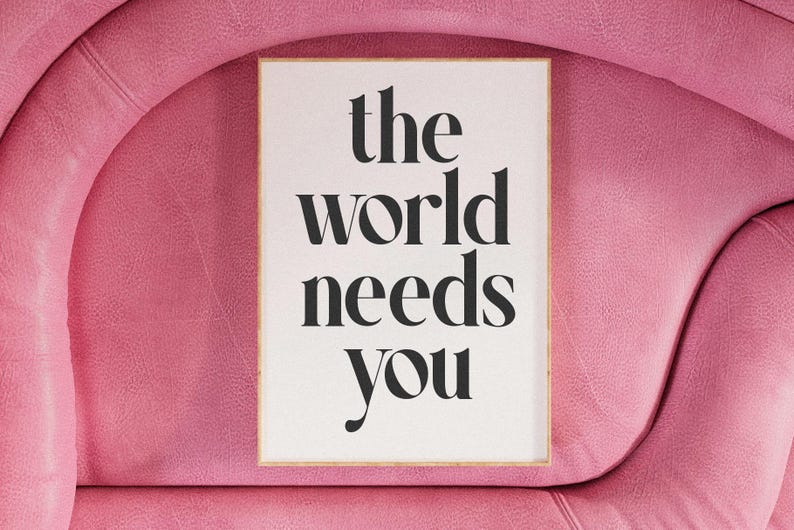 May include: A framed print with the words "the world needs you" in black, elegant typography. The print is set against a white background and framed in a light wood. The background is a pink, textured surface.