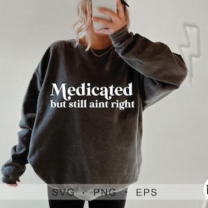 以下が含まれることがあります： 「Medicated but still ain't right」の白い文字が入った濃いグレーのスウェットシャツ。長袖のスウェットシャツはゆったりとしたフィット感です。画像の下部には「SVG . PNG . EPS」という文字と、円の中に「BCD」の文字が表示されています。