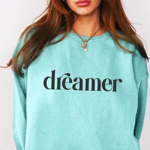May include: A light blue sweatshirt with the word "dreamer" printed in black script. The sweatshirt is a solid color and has a crew neck. The model is wearing a gold necklace.