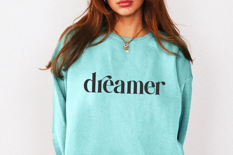 May include: A light blue sweatshirt with the word "dreamer" printed in black script. The sweatshirt is a solid color and has a crew neck. The model is wearing a gold necklace.