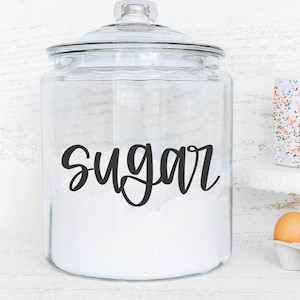 K&ouml;nnte beinhalten: Ein klares Glasgef&auml;&szlig; gef&uuml;llt mit wei&szlig;em Zucker, beschriftet mit dem Wort "sugar" in schwarzer Schrift. Das Gef&auml;&szlig; hat einen Glasdeckel. Ein kleiner Kuchen mit bunten Streuseln und ein Eierkarton befinden sich im Hintergrund.