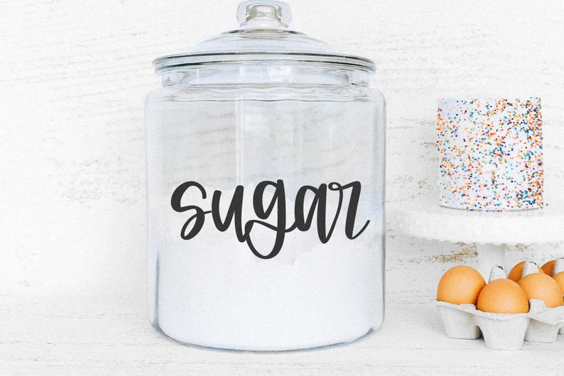 K&ouml;nnte beinhalten: Ein klares Glasgef&auml;&szlig; gef&uuml;llt mit wei&szlig;em Zucker, beschriftet mit dem Wort "sugar" in schwarzer Schrift. Das Gef&auml;&szlig; hat einen Glasdeckel. Ein kleiner Kuchen mit bunten Streuseln und ein Eierkarton befinden sich im Hintergrund.