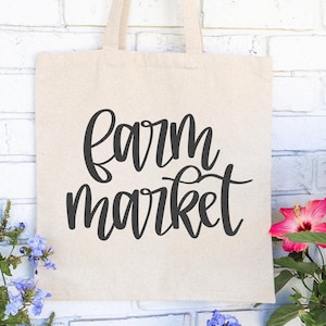 K&ouml;nnte beinhalten: Beige Tragetasche mit dem Schriftzug "Farm Market" in schwarzer Schrift. Die Tasche h&auml;ngt vor einer wei&szlig;en Backsteinmauer, mit rosa und roten Blumen rechts und blauen Blumen links.