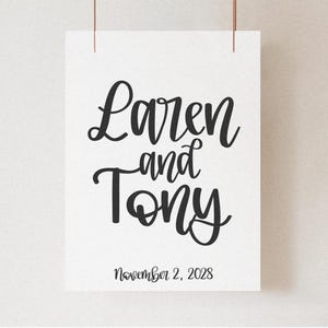 K&ouml;nnte beinhalten: Ein wei&szlig;er Papierdruck mit den Namen "Laren and Tony" in eleganter schwarzer Schrift. Darunter steht das Datum "November 2, 2028". Der Druck wird von zwei d&uuml;nnen, braunen Kordeln vor einem neutralen Hintergrund gehalten.