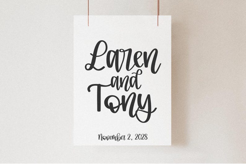 K&ouml;nnte beinhalten: Ein wei&szlig;er Papierdruck mit den Namen "Laren and Tony" in eleganter schwarzer Schrift. Darunter steht das Datum "November 2, 2028". Der Druck wird von zwei d&uuml;nnen, braunen Kordeln vor einem neutralen Hintergrund gehalten.