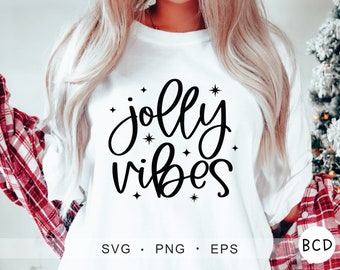 May include: White long-sleeve shirt with black text that reads "jolly vibes" with small stars around the text. The shirt is being worn by a person with long blonde hair. The shirt is being worn with a red and black plaid shirt.