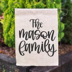 K&ouml;nnte beinhalten: Ein rechteckiger, beigefarbener Gartenflagge mit dem schwarzen Schriftzug "The Mason Family". Die Flagge h&auml;ngt an einem schwarzen Metallst&auml;nder, mit gr&uuml;nem Laub im Hintergrund.