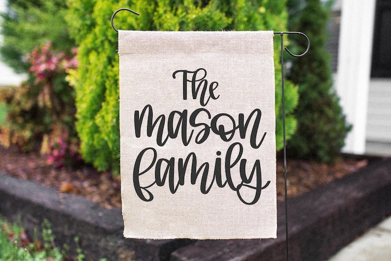 K&ouml;nnte beinhalten: Ein rechteckiger, beigefarbener Gartenflagge mit dem schwarzen Schriftzug "The Mason Family". Die Flagge h&auml;ngt an einem schwarzen Metallst&auml;nder, mit gr&uuml;nem Laub im Hintergrund.