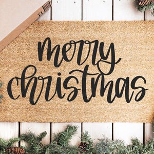 K&ouml;nnte beinhalten: Eine rechteckige Kokos-Fu&szlig;matte mit den Worten "merry christmas" in schwarzer Schrift. Die Matte ist von einem Weihnachtskranz mit Tannennadeln und Tannenzapfen umgeben. Ein Pappkarton befindet sich in der oberen linken Ecke.