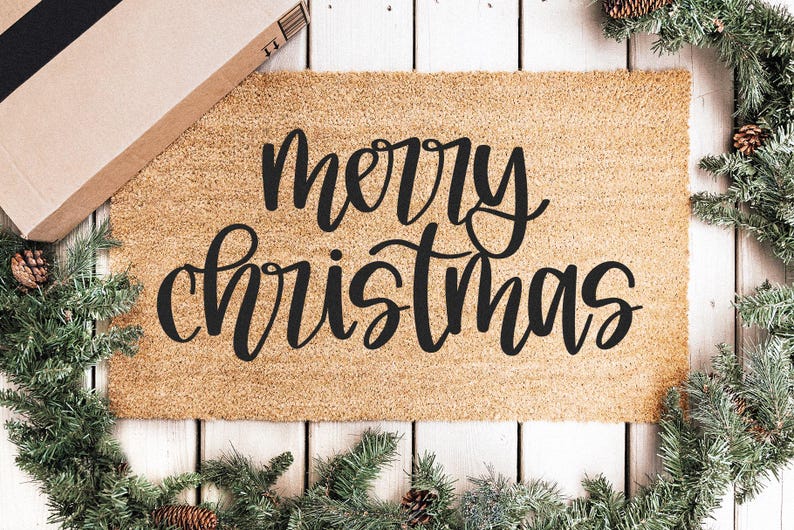 K&ouml;nnte beinhalten: Eine rechteckige Kokos-Fu&szlig;matte mit den Worten "merry christmas" in schwarzer Schrift. Die Matte ist von einem Weihnachtskranz mit Tannennadeln und Tannenzapfen umgeben. Ein Pappkarton befindet sich in der oberen linken Ecke.