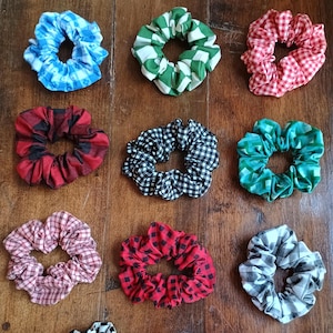 May include: A collection of scrunchies in various patterns and colors, including blue gingham, green and white checkered, red and white gingham, red and black plaid, and black and white checkered. The scrunchies are arranged on a wooden surface.