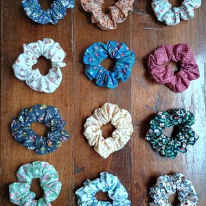 May include: A collection of twelve fabric scrunchies in various colors and floral patterns. The scrunchies are arranged on a wooden surface. Colors include blue, brown, white, teal, pink, navy, yellow, green, and light blue.