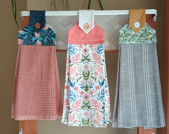 May include: Three hanging kitchen towels with fabric toppers secured by buttons. The left towel is solid dusty rose with a dark teal floral topper. The center towel features a pink and blue floral print. The right towel is grey with white stripes.
