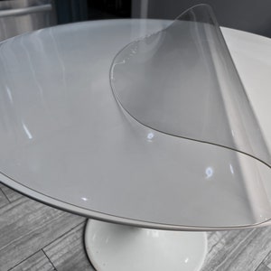 Peut inclure: Une table ronde blanche avec une nappe en plastique transparente. La nappe est légèrement froissée et a un bord incurvé.