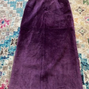 May include: A long, purple suede skirt with a straight cut and a simple design. The skirt has a zipper closure and two pockets on the front.