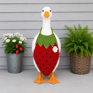 May include: A white goose figurine wearing a crocheted strawberry-shaped garment. The garment features a green top section, a red body with small black embroidered seeds, and a single white flower accent on the side. The goose has orange feet.