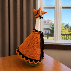 May include: A white goose figurine wearing a crocheted orange and black cape with a scalloped edge and a matching orange and black pointed hat. The cape features a black collar and black ties, while the hat is secured with a thin black ribbon.
