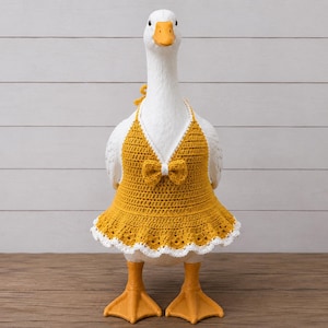 Può includere: Una statuetta di oca bianca che indossa un abito all'americana all'uncinetto color giallo senape. L'abito presenta un piccolo fiocco al centro del petto e un bordo in pizzo bianco smerlato lungo l'orlo inferiore. L'oca ha becco e zampe arancioni.