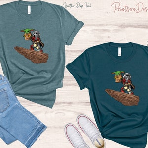 May include: Two teal t-shirts with a cartoon image of a Mandalorian character carrying a small green alien. The text "Printson Designs" is printed on the top of the image.