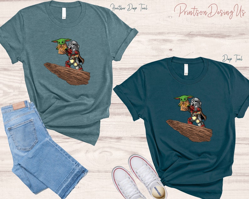 May include: Two teal t-shirts with a cartoon image of a Mandalorian character carrying a small green alien. The text "Printson Designs" is printed on the top of the image.