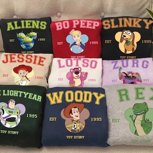 Puede incluir: Nueve sudaderas con dise&ntilde;os de personajes de "Toy Story". Cada sudadera presenta nombres como "Aliens", "Bo Peep" y "Slinky", junto con ilustraciones de personajes y el texto "EST 1995 TOY STORY."
