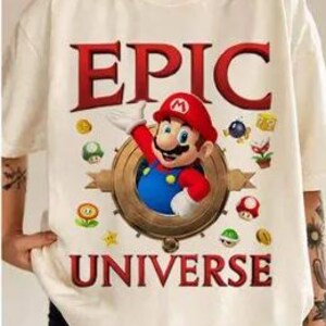 May include: White t-shirt with a graphic of Mario from the Super Mario Bros. video game series. The graphic includes the text "EPIC UNIVERSE" and features Mario in a circular frame with various power-ups and coins.