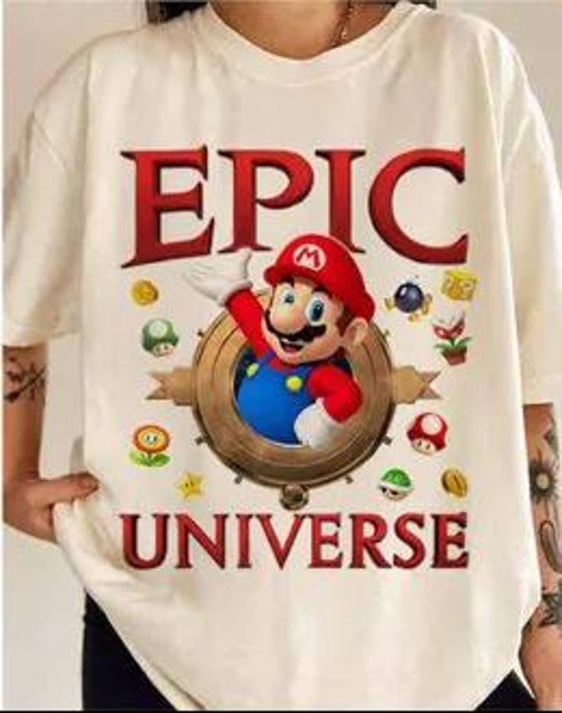 May include: White t-shirt with a graphic of Mario from the Super Mario Bros. video game series. The graphic includes the text "EPIC UNIVERSE" and features Mario in a circular frame with various power-ups and coins.